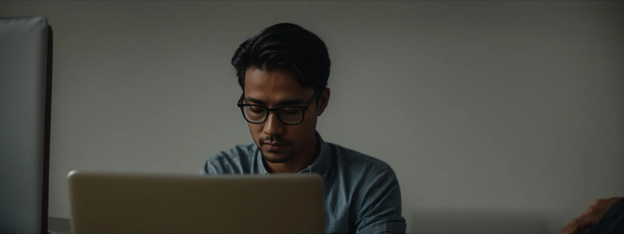 a person thoughtfully typing on a laptop with an open notebook and glasses beside them.