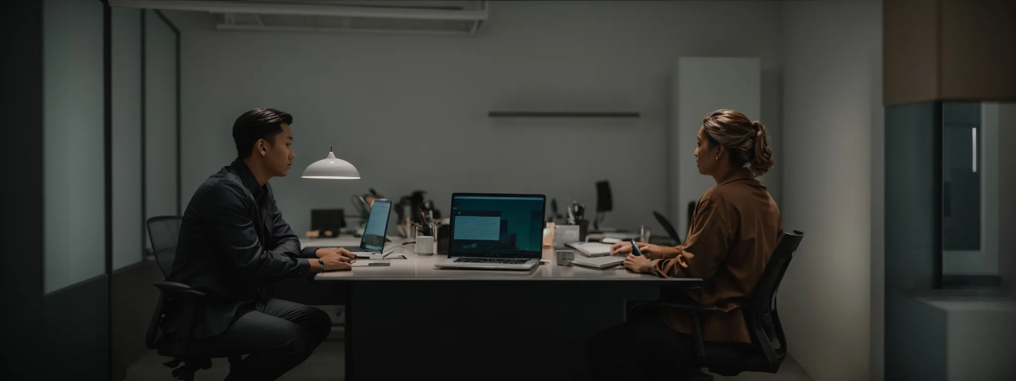 two professionals sit across from each other at a minimalist office desk, each intently focused on a laptop screen while discussing strategy.