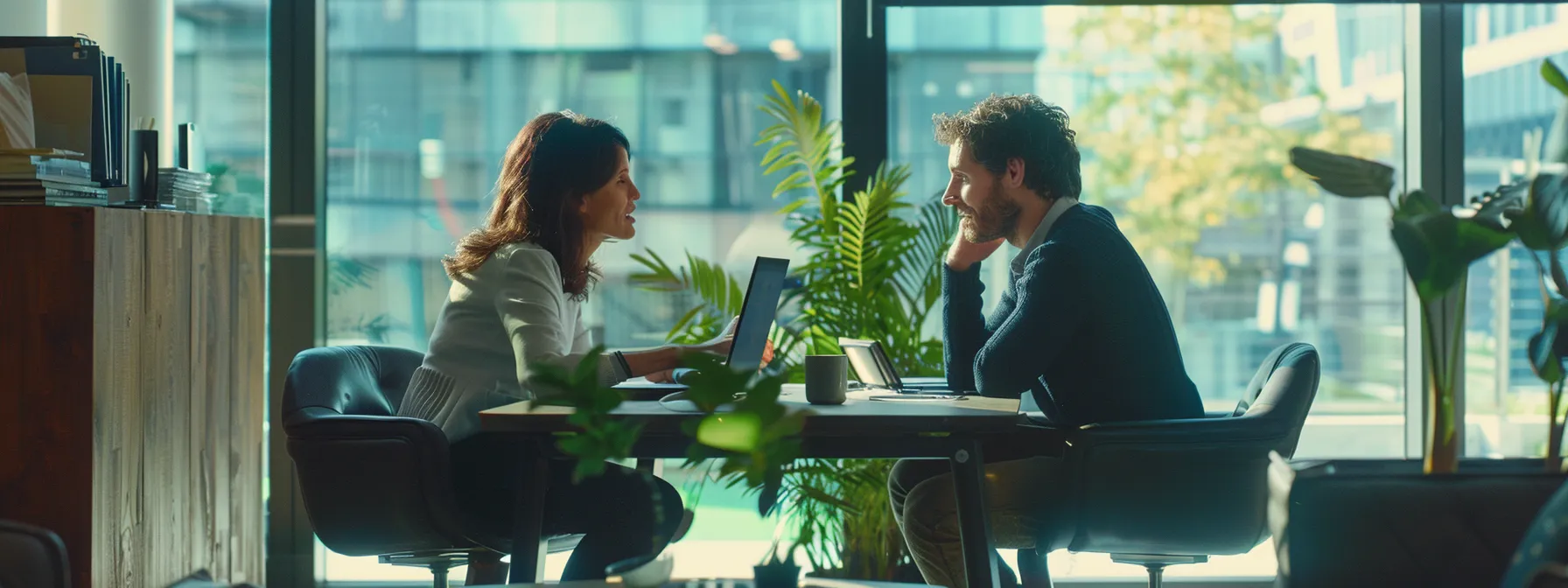 Two people sit across from each other at a table in a well-lit office, engaged in a conversation about holistic SEO strategies. A laptop and documents are on the table, with plants and large windows in the background.