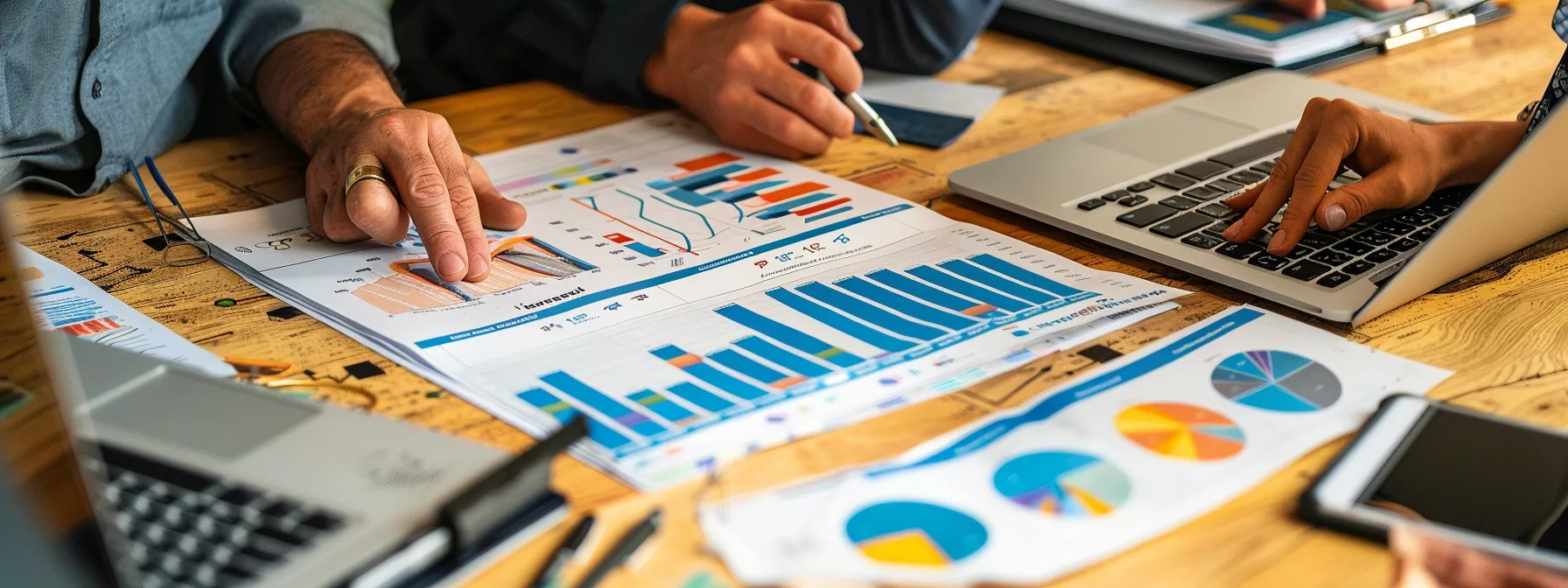 Hands of individuals working with laptops and analyzing colorful graphs and charts on paper spread out on a wooden desk, boosting site performance through comprehensive analysis.